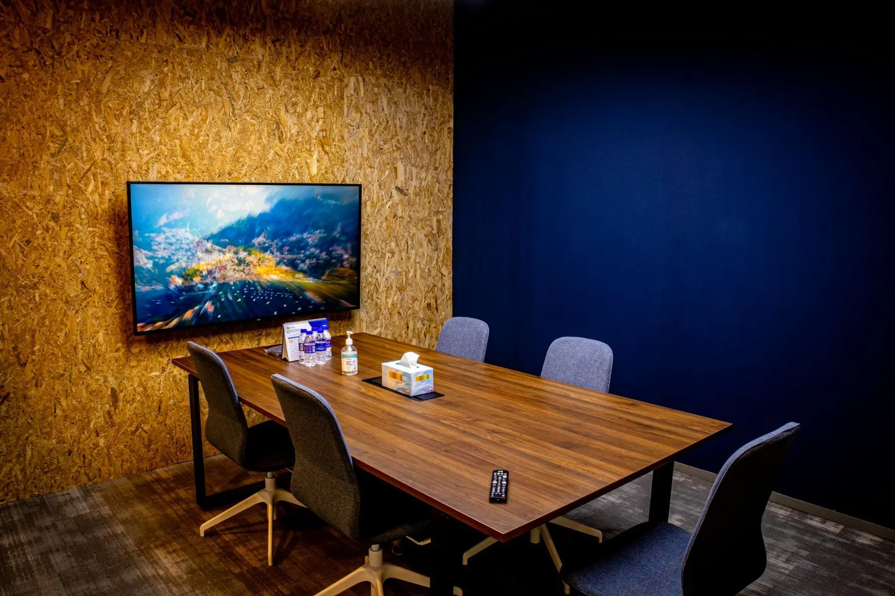 A meeting room with a wooden conference table, chairs and a wall-mounted flat-screen display.