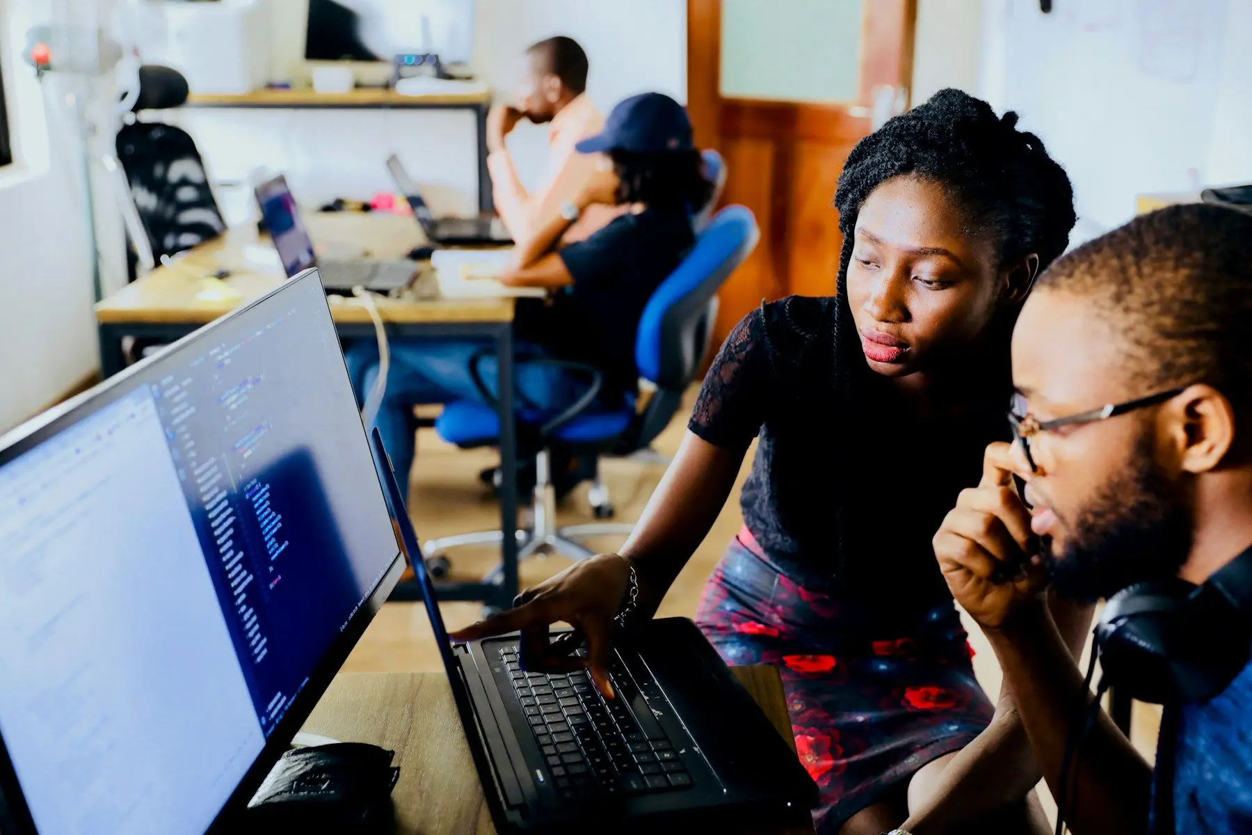 Two colleagues leaning in together to look at a laptop screen in a busy office.