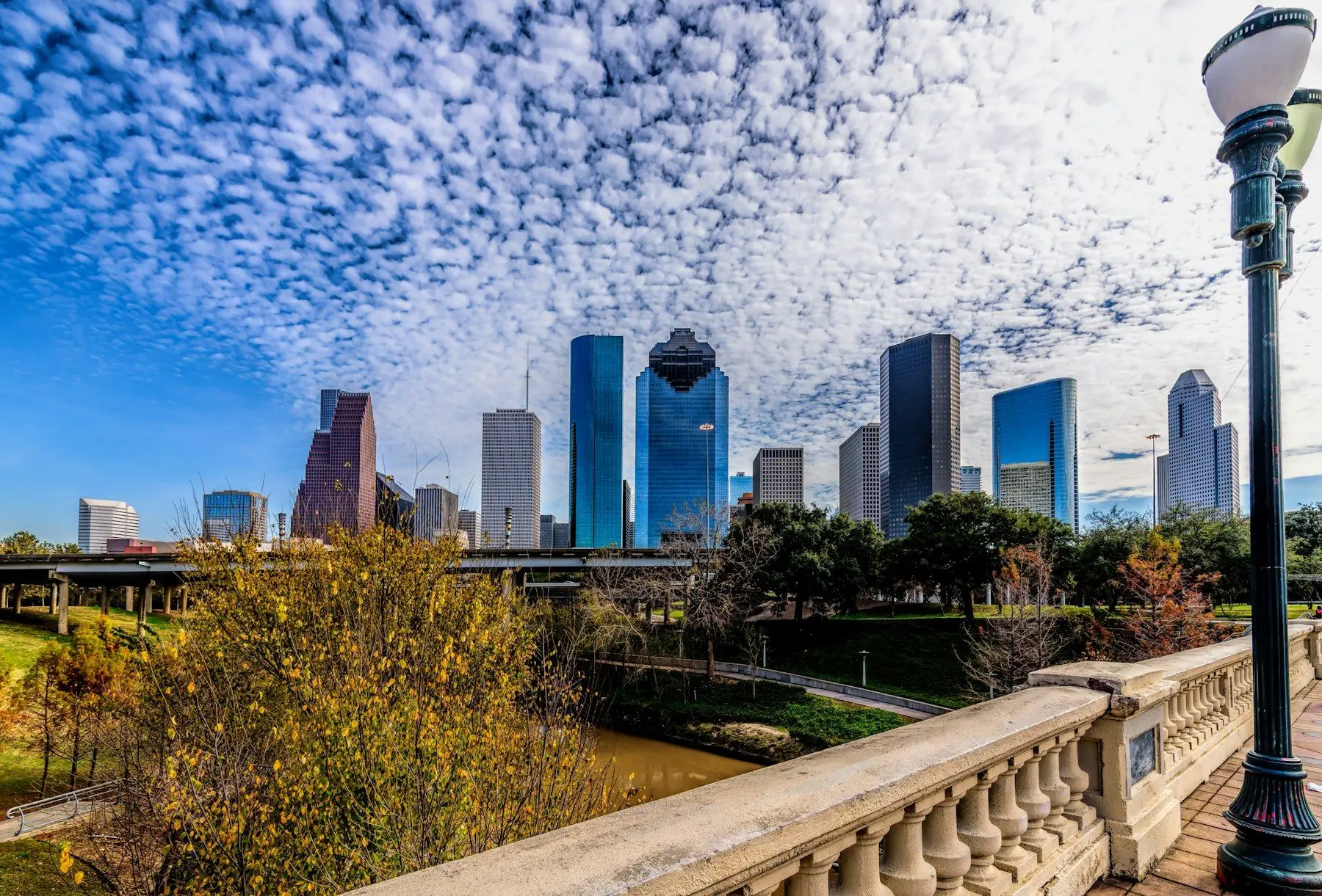 The downtown Houston skyline on a sunny day, seen past trees and a stone bridge railing.